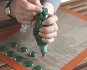 Hands squeeze a green icing piping bag to pipe round green dollops on a silicone baking mat.