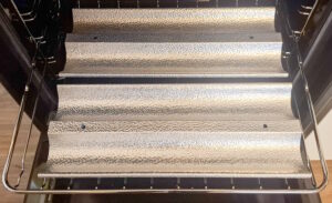 Inside an oven, a chrome pull-out rack holding three textured metallic baking trays lined up
