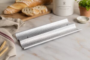 Two textured silver aluminum foil strips laid on a marble kitchen counter, with bread, a linen cloth, and a plant in the background.