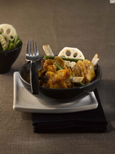 Bowl of braised chicken with vegetables and lotus root slices, on a white square plate with a fork nearby.