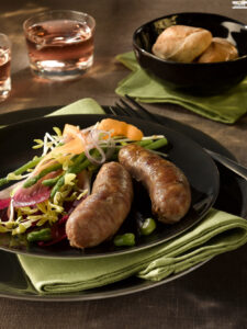Two sausages on a black plate with a fresh salad (sprouts, greens, onions) and a side of bread in a black bowl in the background.
