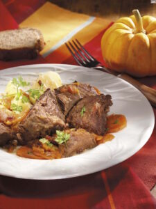 Plate of braised beef with potatoes and carrot gravy, garnished with parsley, on a white plate with autumn decor (pumpkin and bread) nearby.