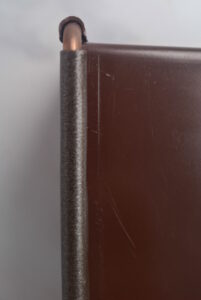 Close-up of a copper pin and gray metal post mounted at the edge of a dark brown panel with a small cap on top.