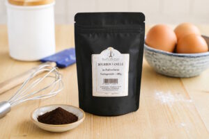 Black pouch labeled Bourbon Vanille in Pulverform on a kitchen table, with a small dish of brown powder in front and eggs in a bowl to the right.