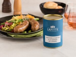 Blue Lafitte can labeled 'Saucisses confites à la graisse de canard' next to a plated sausage dish with salad greens and bread in the background.